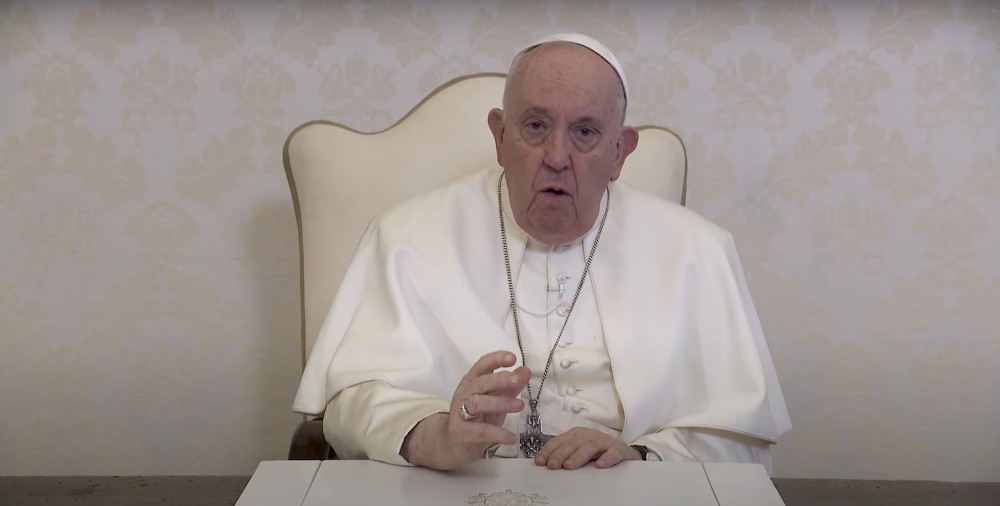 Pope Francis sits in a white backed chair against a white patterned wallpaper wall and speaks