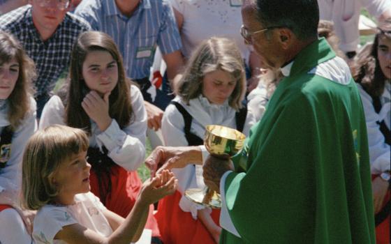 A man in green vestments distributes Communion to a group of white people outside